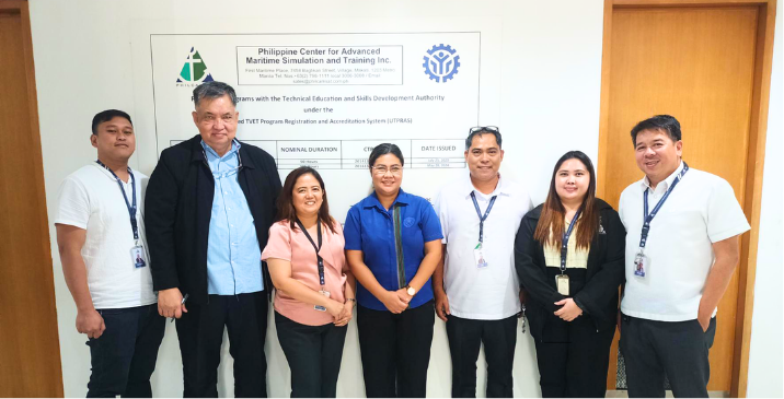 Seven staff members with lanyards pose in a hallway in front of a sign for a maritime training center.